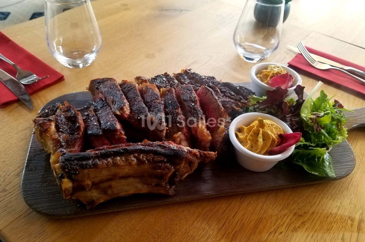 Planche de côtes de bœuf grillées accompagnée de salade verte et de deux ramequins de sauce sur une table en bois.