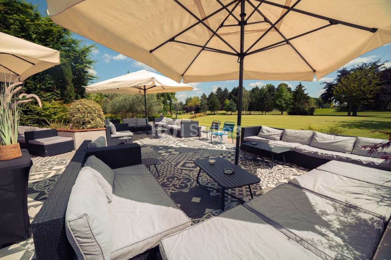 Terrasse aménagée avec canapés, tables basses et parasols, donnant sur un grand jardin verdoyant.