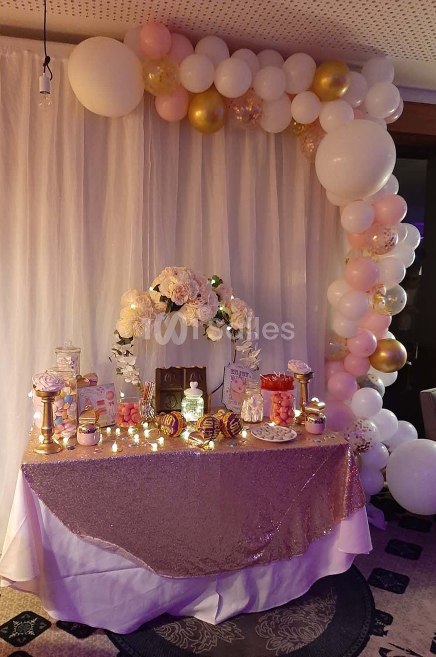 Table décorée pour une fête avec nappes scintillantes, fleurs, ballons blancs et dorés, et desserts variés.