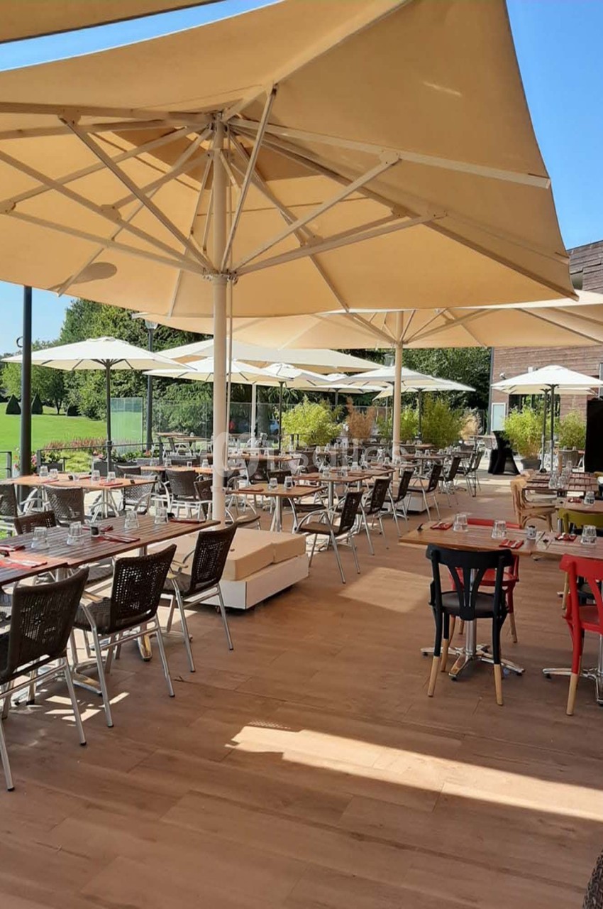 Terrasse d'un restaurant avec tables dressées, chaises et grands parasols, donnant sur un espace vert par temps ensoleillé.