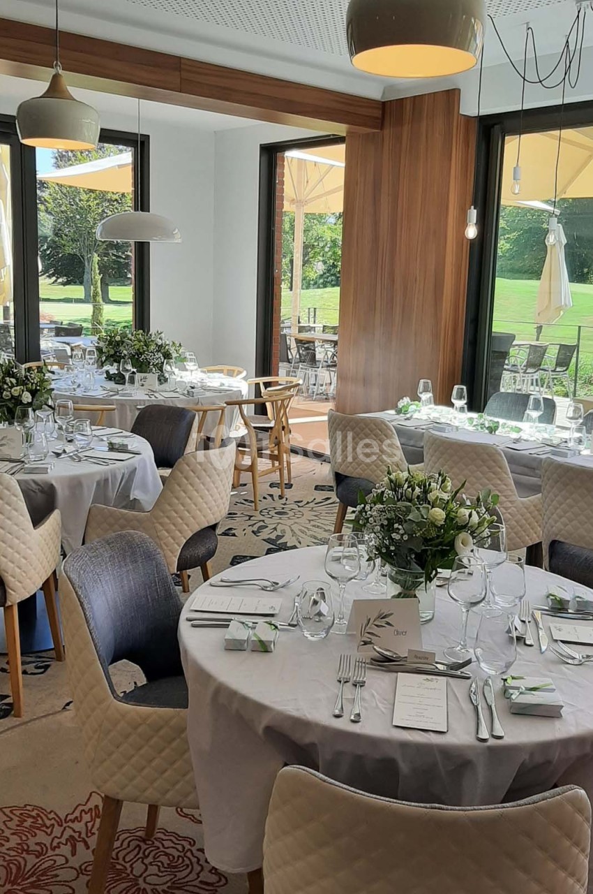 Salle de réception lumineuse avec tables rondes dressées, décorées de fleurs blanches, donnant sur une terrasse et un jardin.