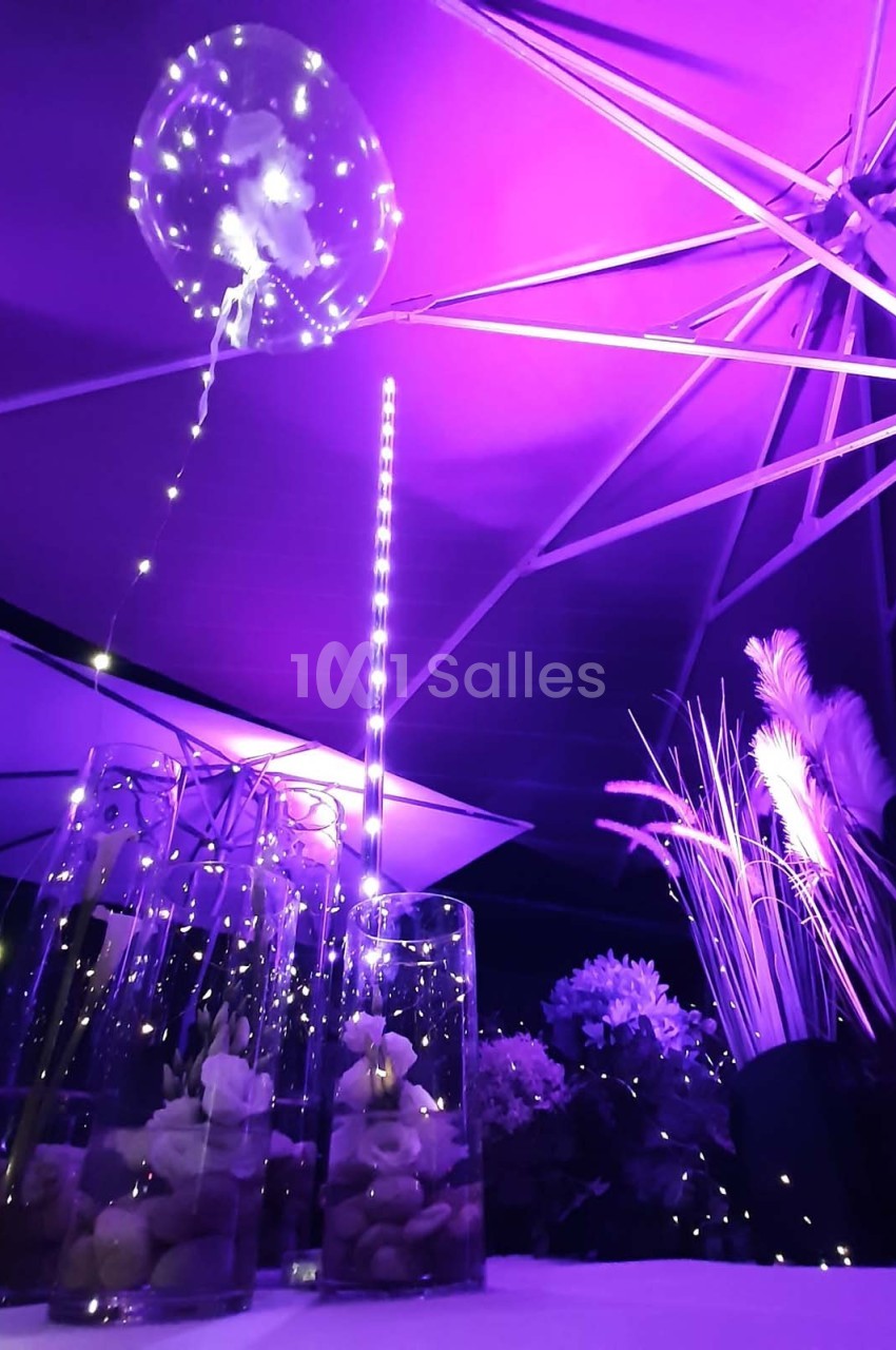 Décoration nocturne avec guirlandes lumineuses, parasols éclairés en violet et vases contenant des galets et des fleurs.