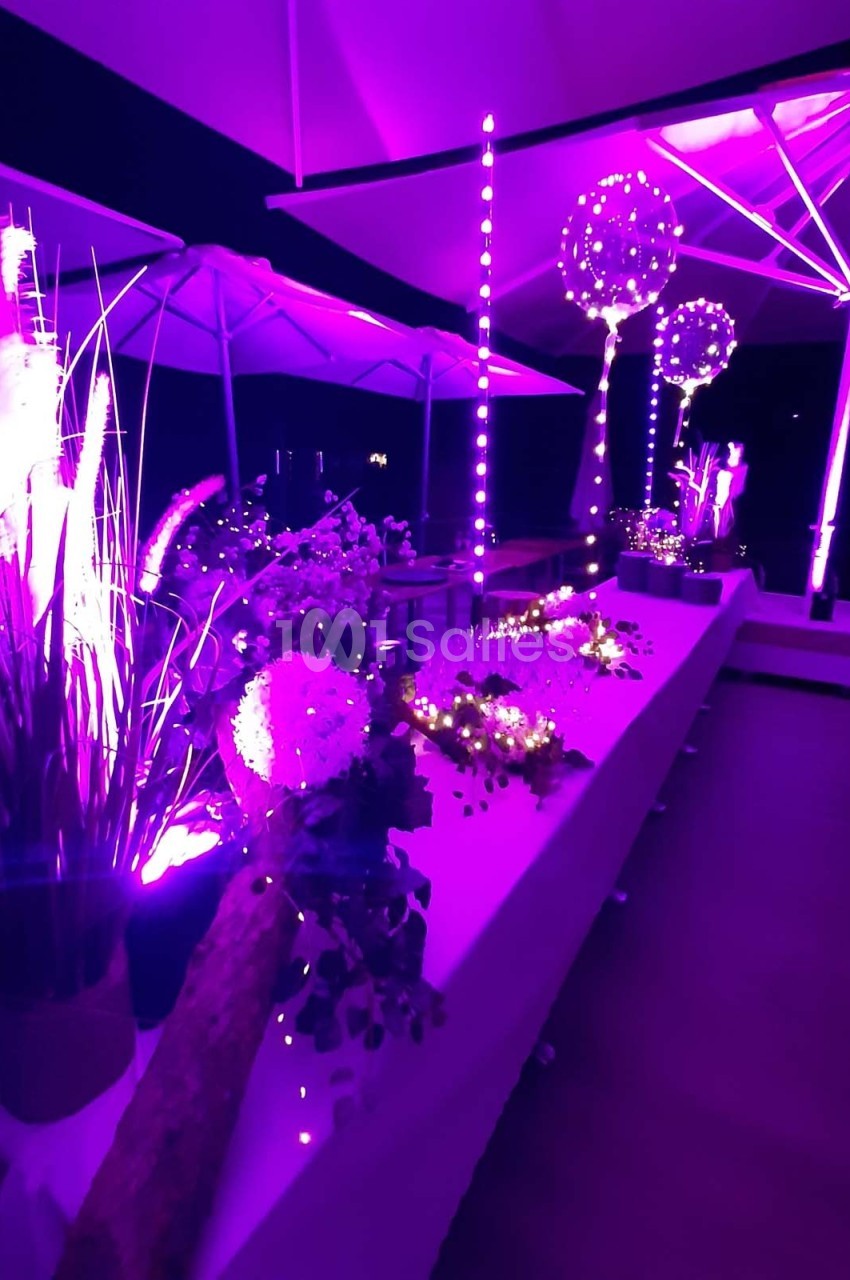 Décoration lumineuse violette avec fleurs, guirlandes et parasols éclairés dans un espace intérieur sombre.