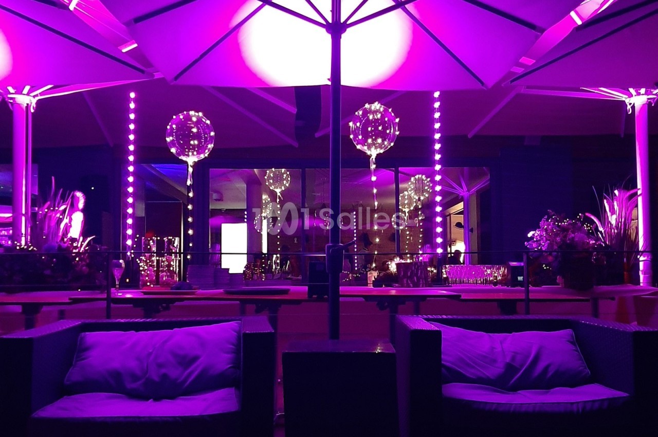 Terrasse extérieure éclairée par des lumières violettes, avec parasols, fauteuils et décoration lumineuse.