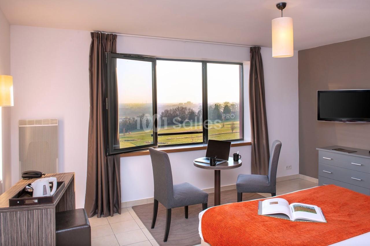 Chambre lumineuse avec lit, table ronde, deux chaises, fenêtre donnant sur un paysage, et télévision murale.