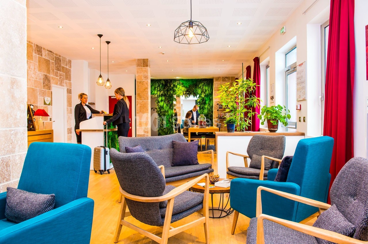 Salon lumineux avec fauteuils colorés, plantes vertes, et deux personnes discutant près d'un comptoir d'accueil.
