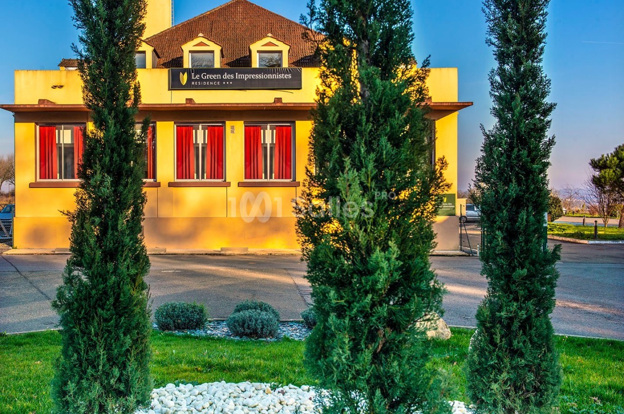 Façade d'un bâtiment jaune avec des volets rouges, entouré de cyprès et d'un parterre de pierres blanches.