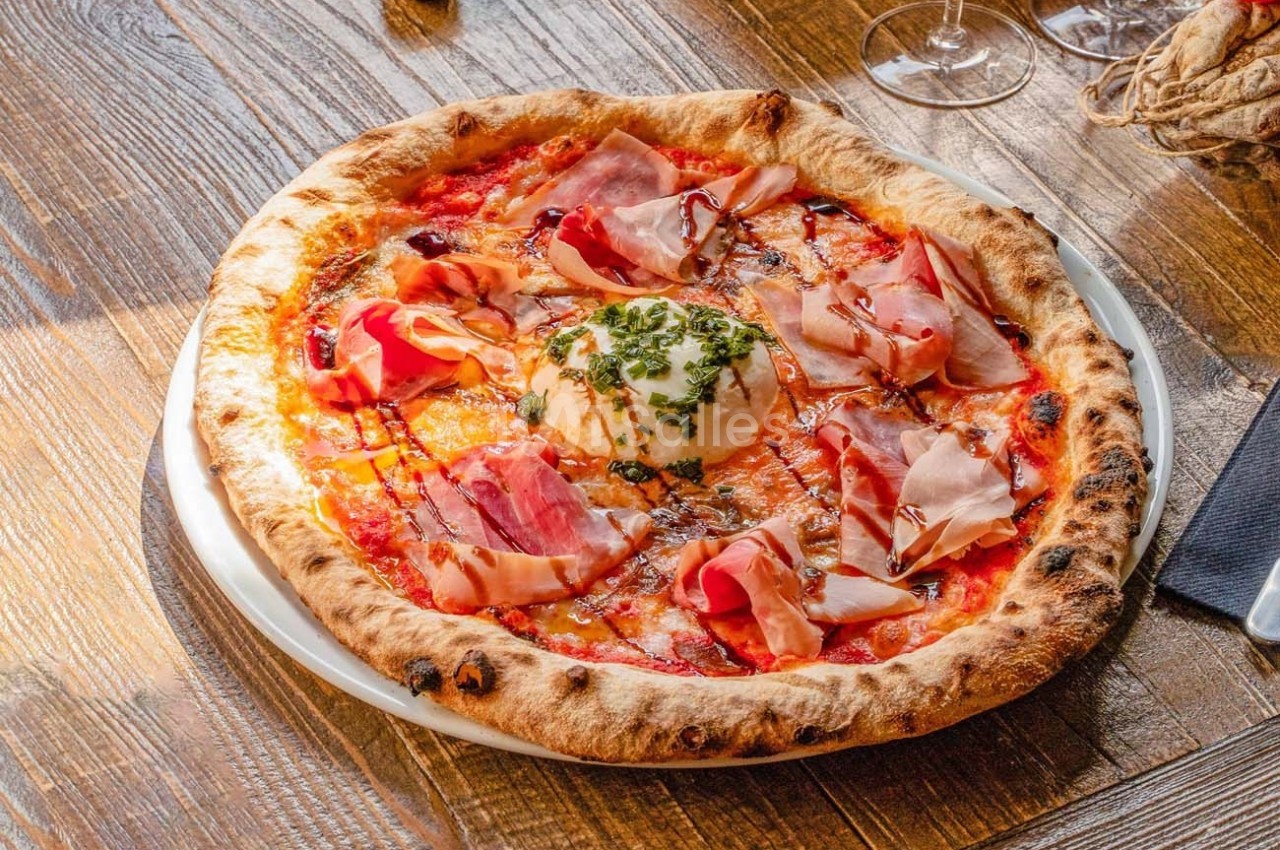 Pizza garnie de jambon cru, burrata au centre, filet de balsamique, servie sur une assiette blanche sur table en bois.