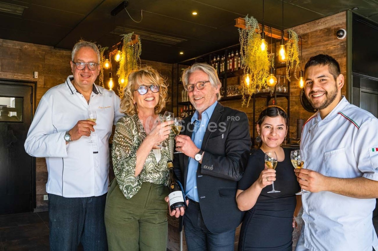 Cinq personnes souriantes trinquent avec des verres de vin dans un intérieur chaleureux décoré de plantes suspendues.