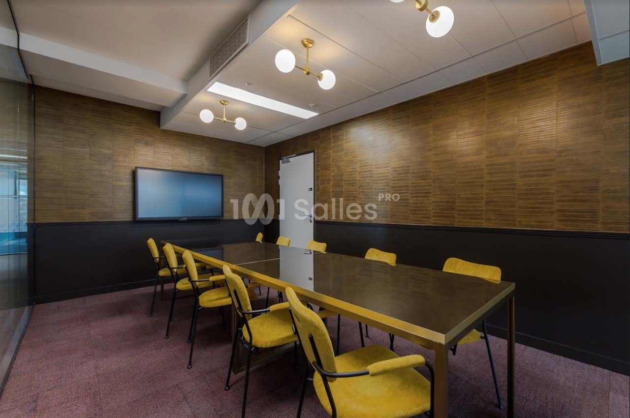 Salle de réunion avec table rectangulaire, chaises jaunes, écran mural et éclairage suspendu.