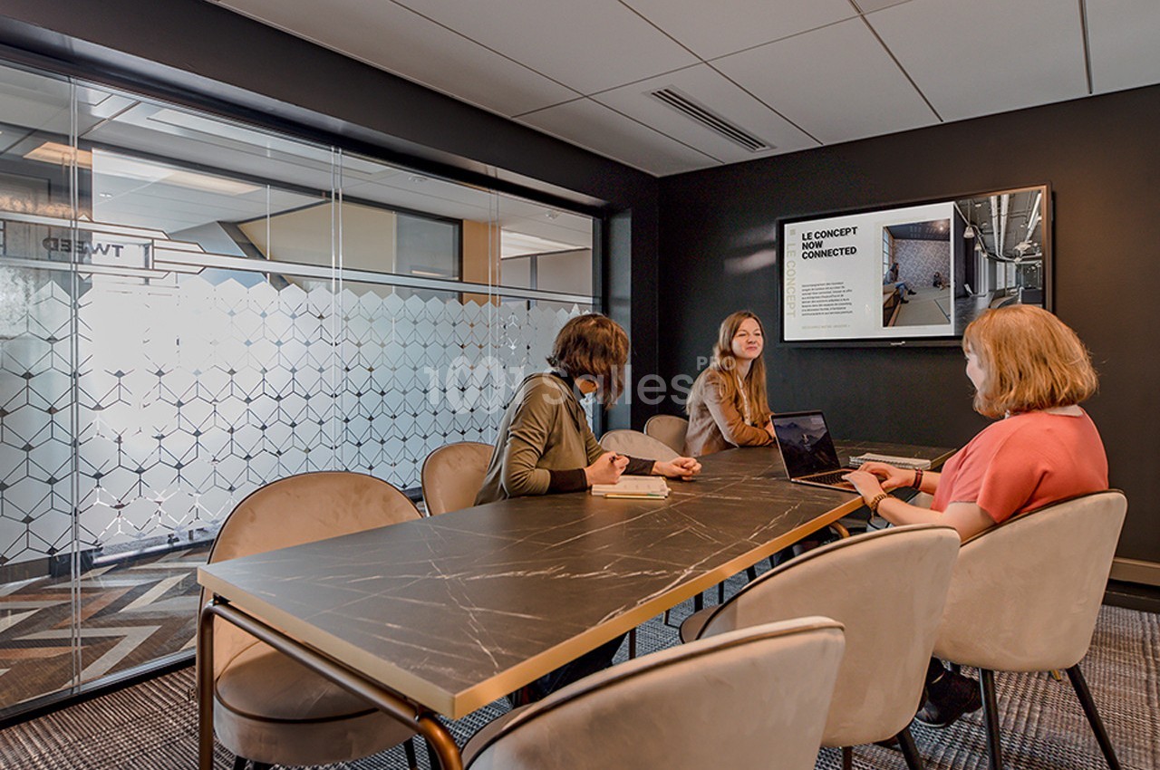 Trois personnes discutent autour d'une table dans une salle de réunion moderne avec un écran affichant une présentation.