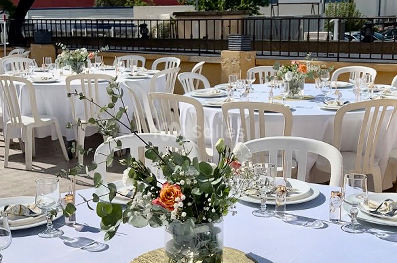 Tables dressées en extérieur avec nappes blanches, chaises en plastique et centres de table fleuris.