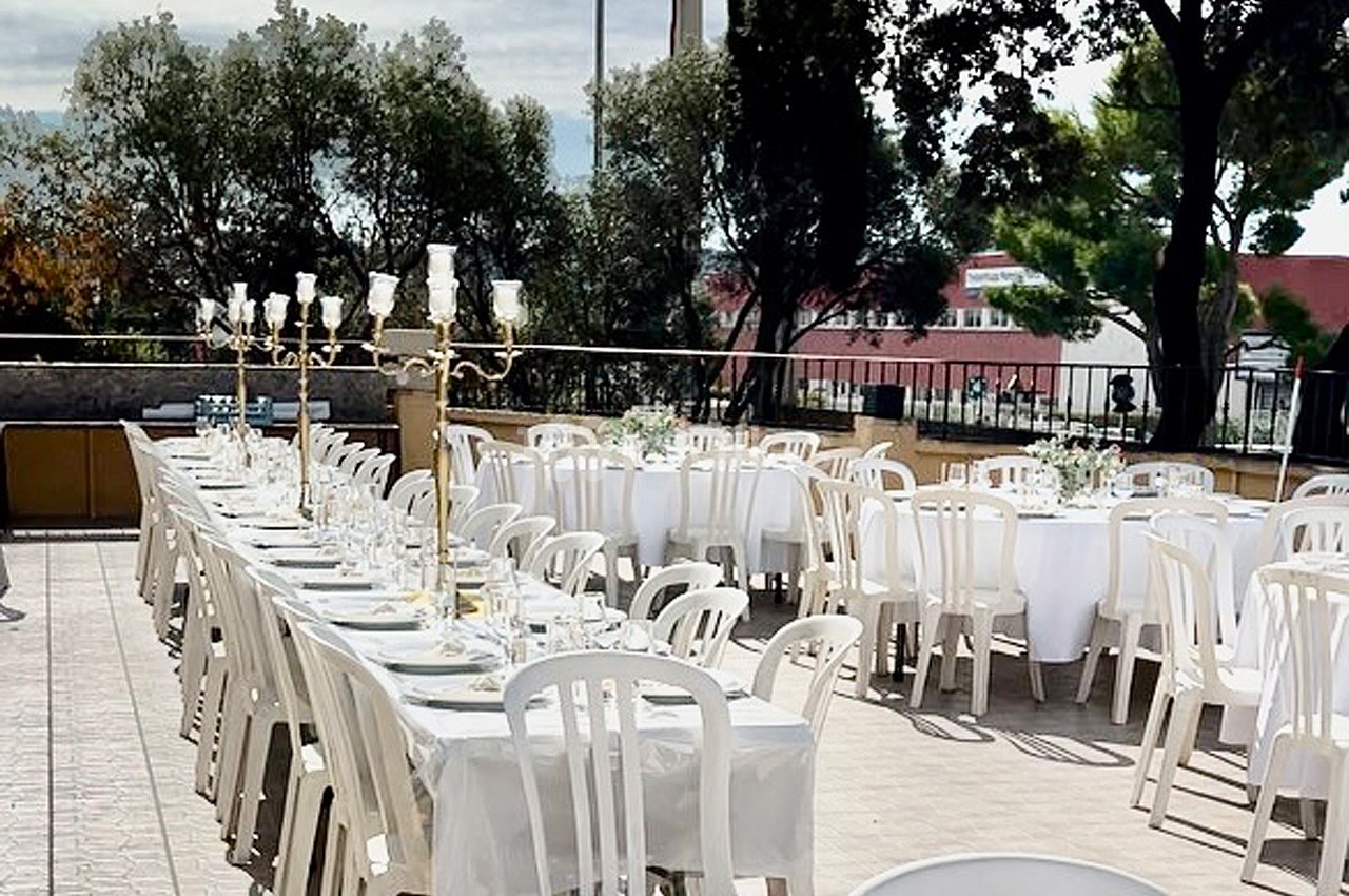 Tables dressées en extérieur avec nappes blanches, chaises en plastique et chandeliers, entourées d'arbres.