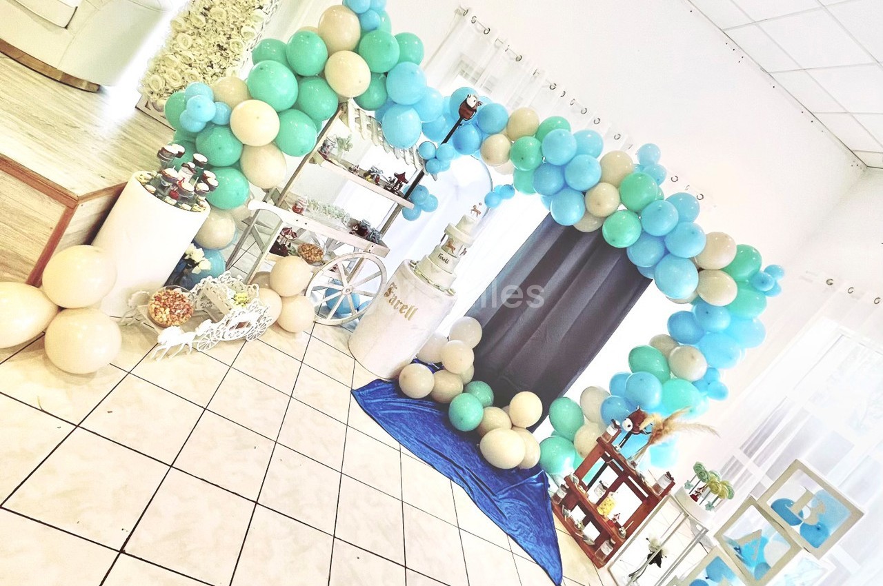 Décoration festive avec arche de ballons bleus et beiges, table de desserts et gâteau central dans une salle lumineuse.