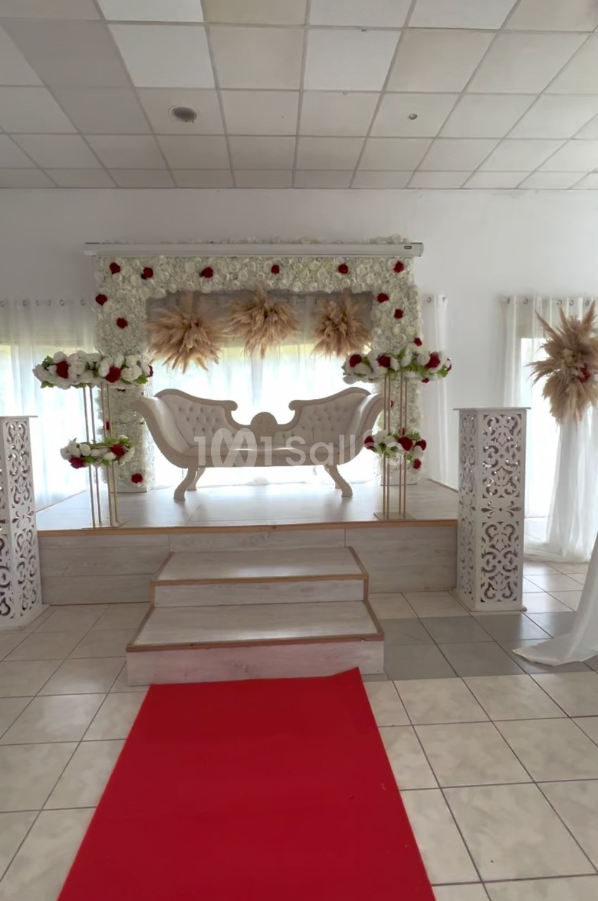 Salle décorée pour un événement avec un canapé blanc sur une estrade, fleurs rouges et blanches, et tapis rouge au sol.