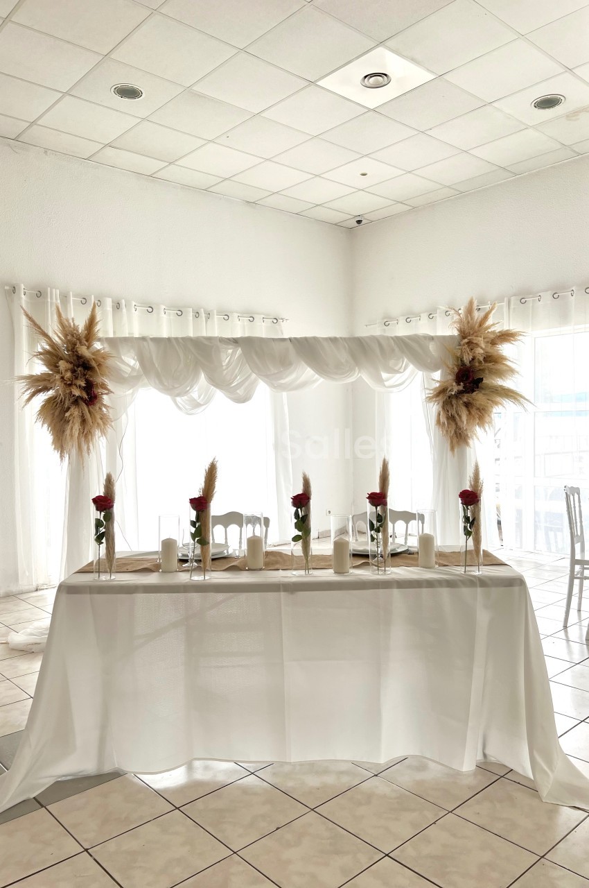 Table décorée pour un événement avec nappes blanches, bougies, fleurs et ornements de pampas dans une salle lumineuse.