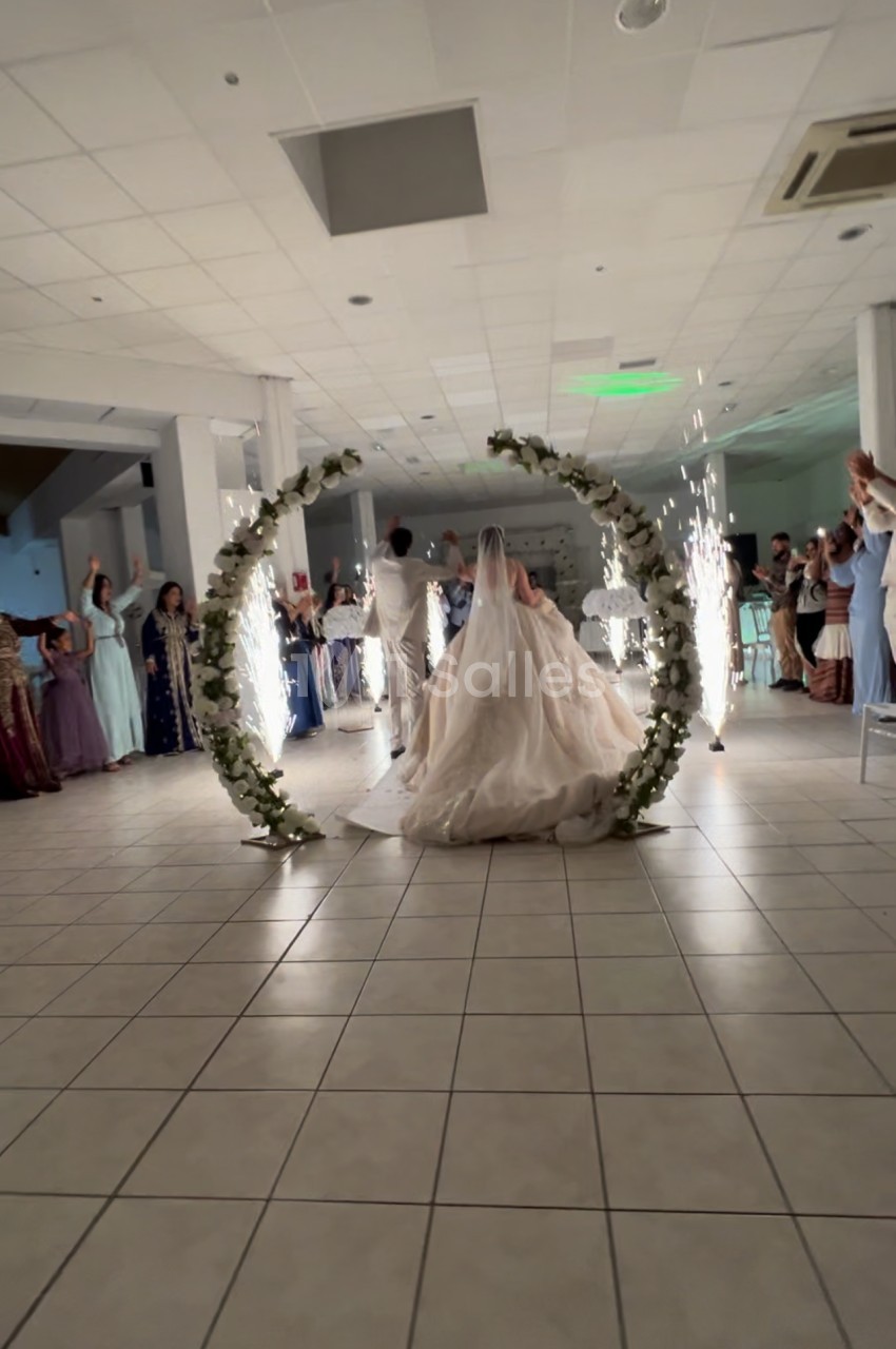 Un couple de mariés traverse une arche fleurie illuminée par des étincelles, entouré d'invités dans une salle de réception.