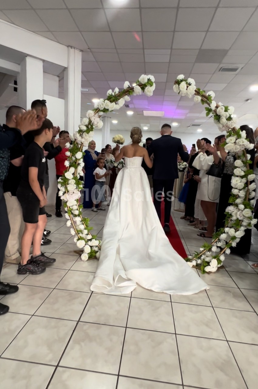 Une mariée en robe blanche et son accompagnateur traversent une arche fleurie devant des invités dans une salle.