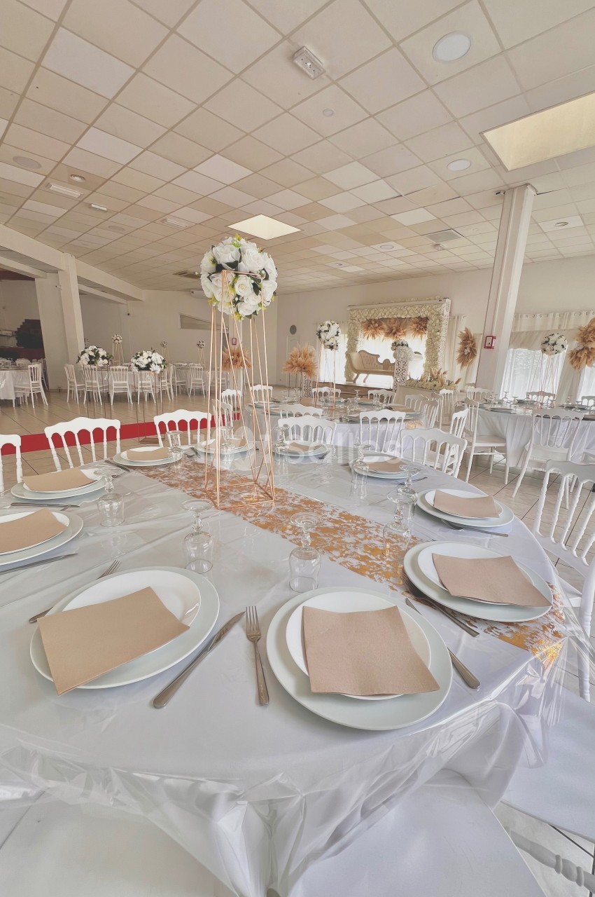 Salle de réception décorée avec des tables dressées, nappes blanches, serviettes beige et centres de table floraux.