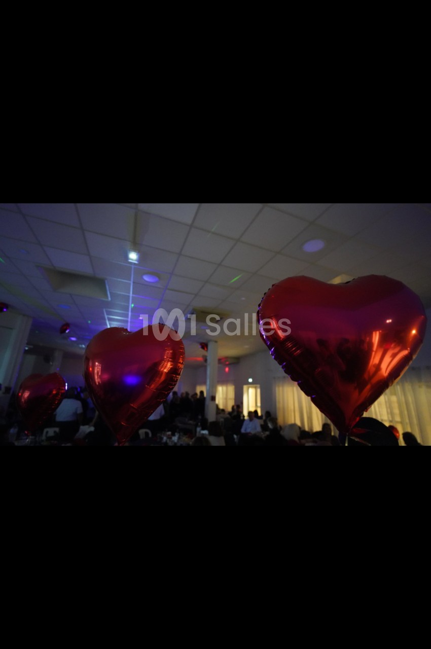Des ballons rouges en forme de cœur flottent dans une salle éclairée, décorée pour un événement festif.