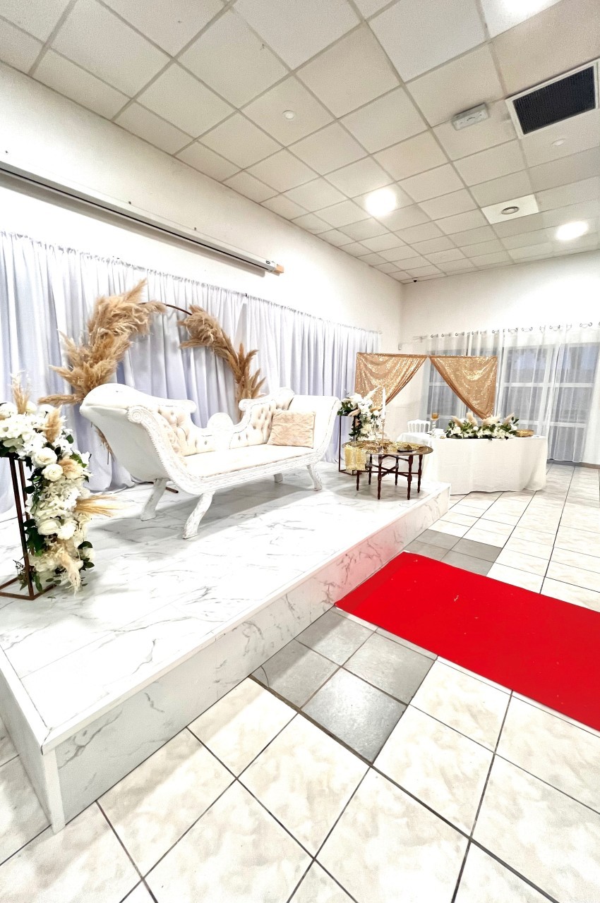 Salle décorée pour un événement avec canapé blanc sur estrade, fleurs, rideaux blancs et tapis rouge au sol.