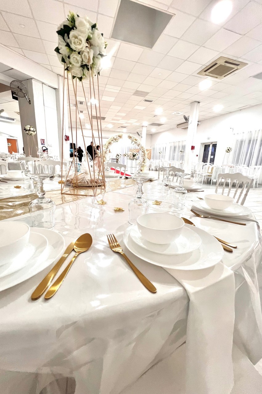 Table de réception élégamment dressée avec vaisselle blanche, couverts dorés et décoration florale dans une salle lumineuse.