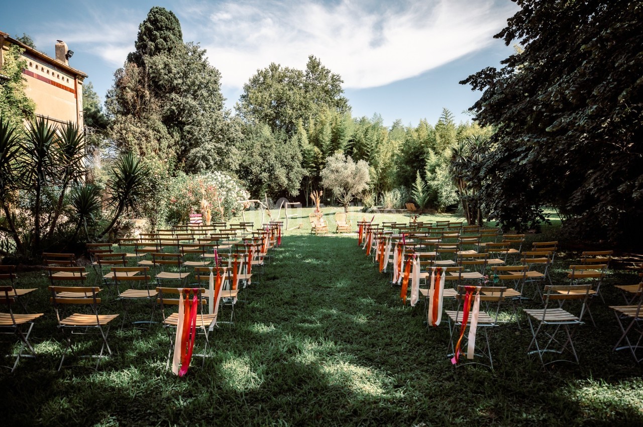 Chaises disposées en rangées sur une pelouse, formant une allée centrale, dans un jardin arboré sous un ciel dégagé.