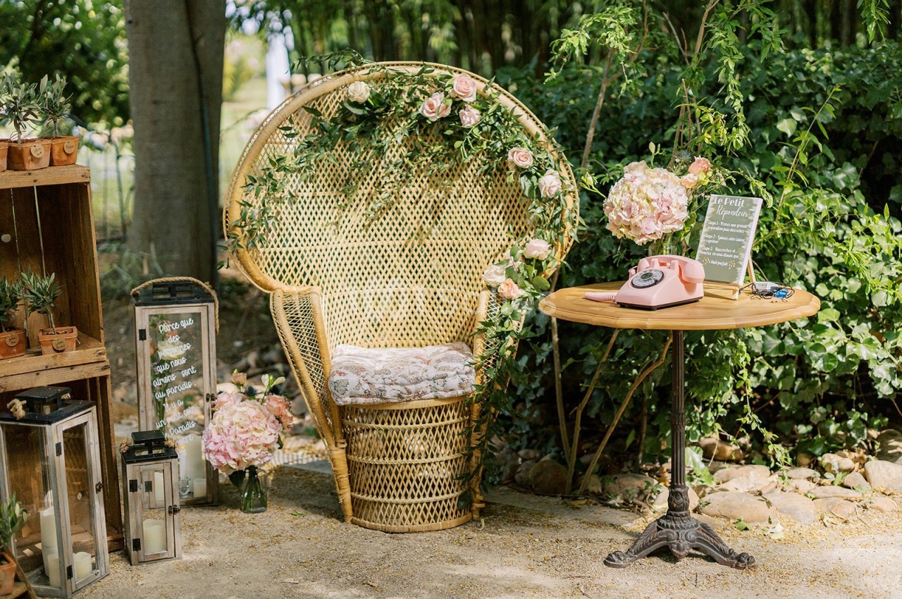 Fauteuil en osier décoré de fleurs, table ronde avec téléphone rose et panneau, entourés de végétation et de lanternes.