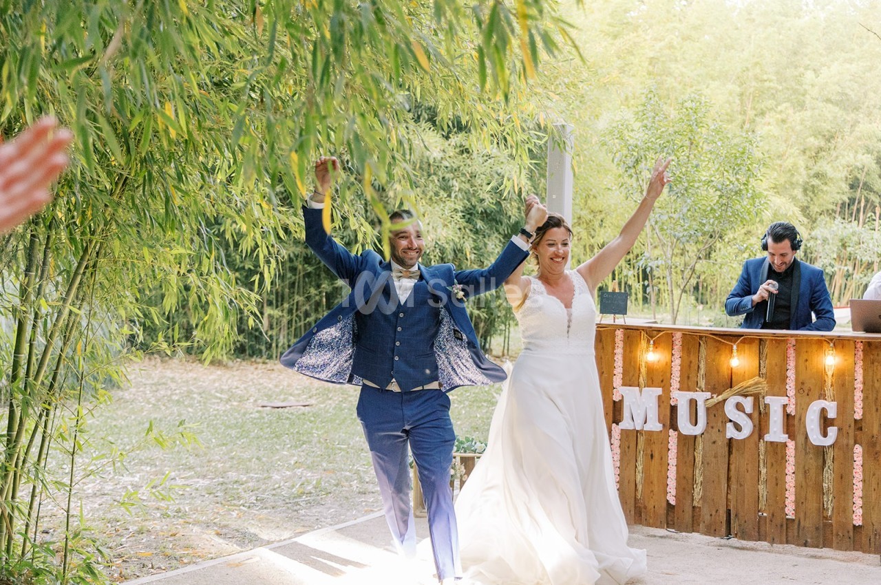 Un couple en tenue de mariage entre sur une piste de danse en plein air, entouré de verdure et d'une décoration musicale.