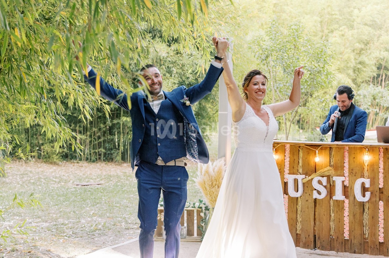Un couple en tenue de mariage lève les bras en souriant, devant un décor champêtre avec un DJ en arrière-plan.