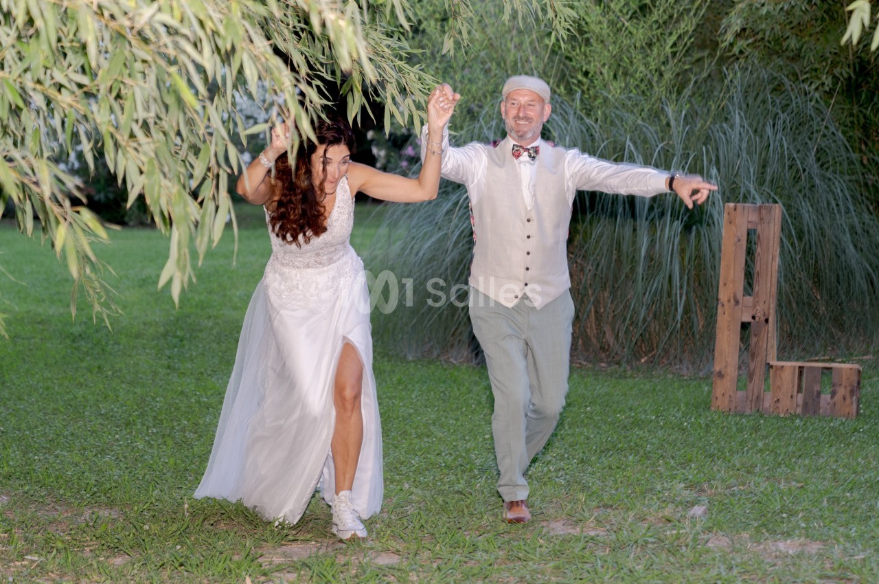 Un couple en tenue de mariage danse joyeusement sur une pelouse entourée de végétation.