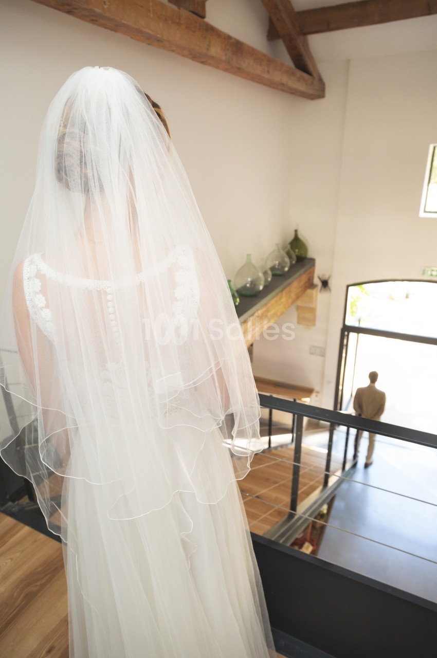Une mariée en robe blanche et voile descend un escalier en bois, regardant un homme debout en bas.