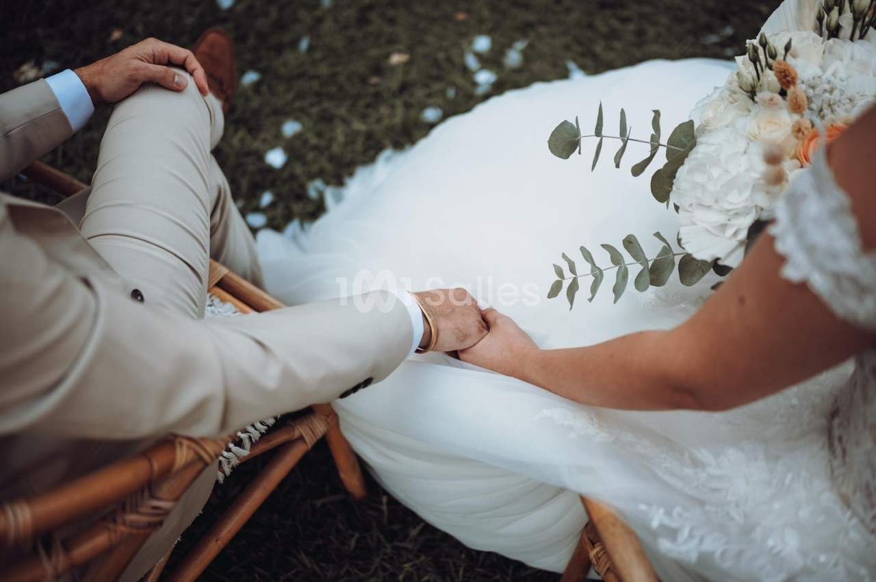Un couple assis se tenant la main, la mariée en robe blanche tenant un bouquet avec des feuilles d'eucalyptus.