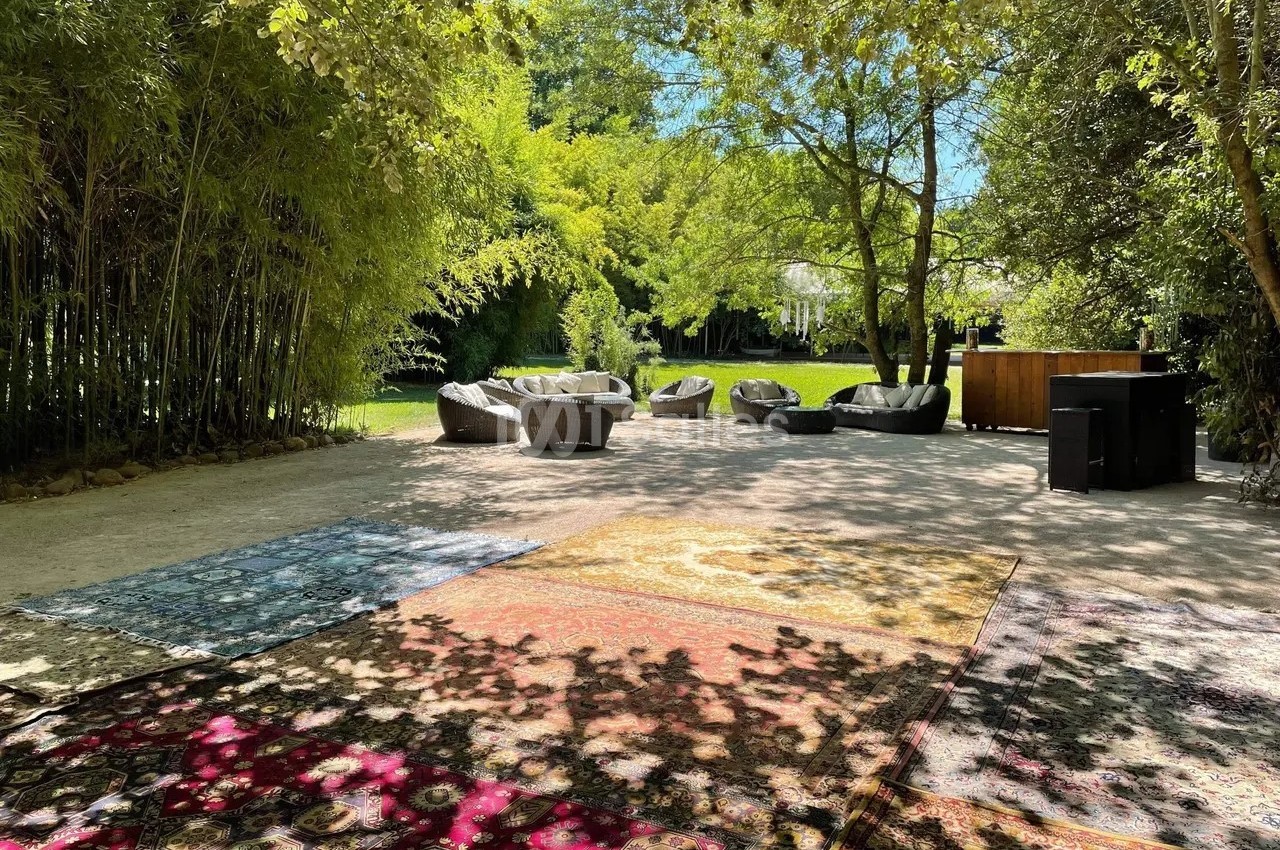 Espace extérieur ombragé avec tapis colorés au sol, fauteuils et végétation luxuriante en arrière-plan.