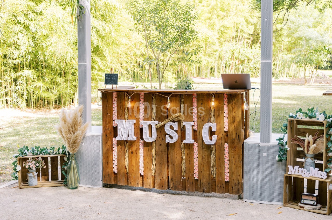 Bar en bois décoré avec des guirlandes lumineuses, des lettres ’MUSIC’ et des éléments végétaux, dans un cadre extérieur.