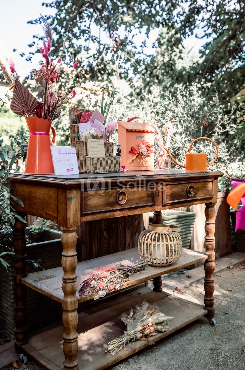 Table en bois avec décorations florales, cartes, arrosoir orange et bouquets séchés, placée dans un jardin ensoleillé.