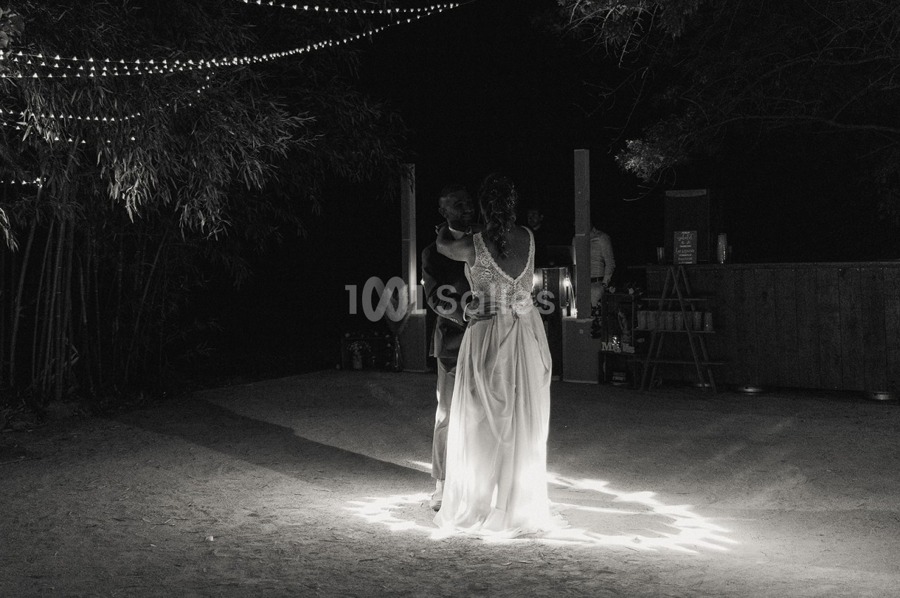 Une mariée en robe blanche se tient sous des guirlandes lumineuses dans un espace extérieur sombre.