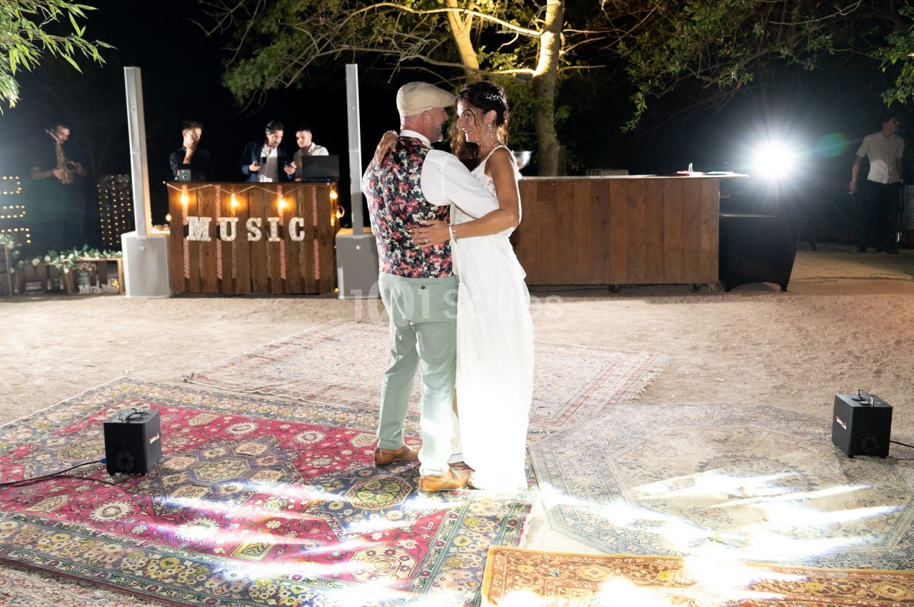 Un couple danse sur des tapis colorés en extérieur, éclairé par des lumières, près d'un bar en bois et d'un panneau ’MUSIC’.
