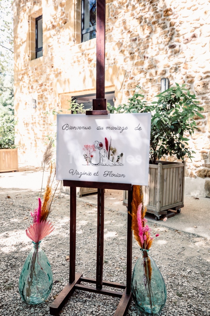 Panneau de bienvenue à un mariage, décoré de fleurs séchées, placé devant un mur en pierre et des plantes.