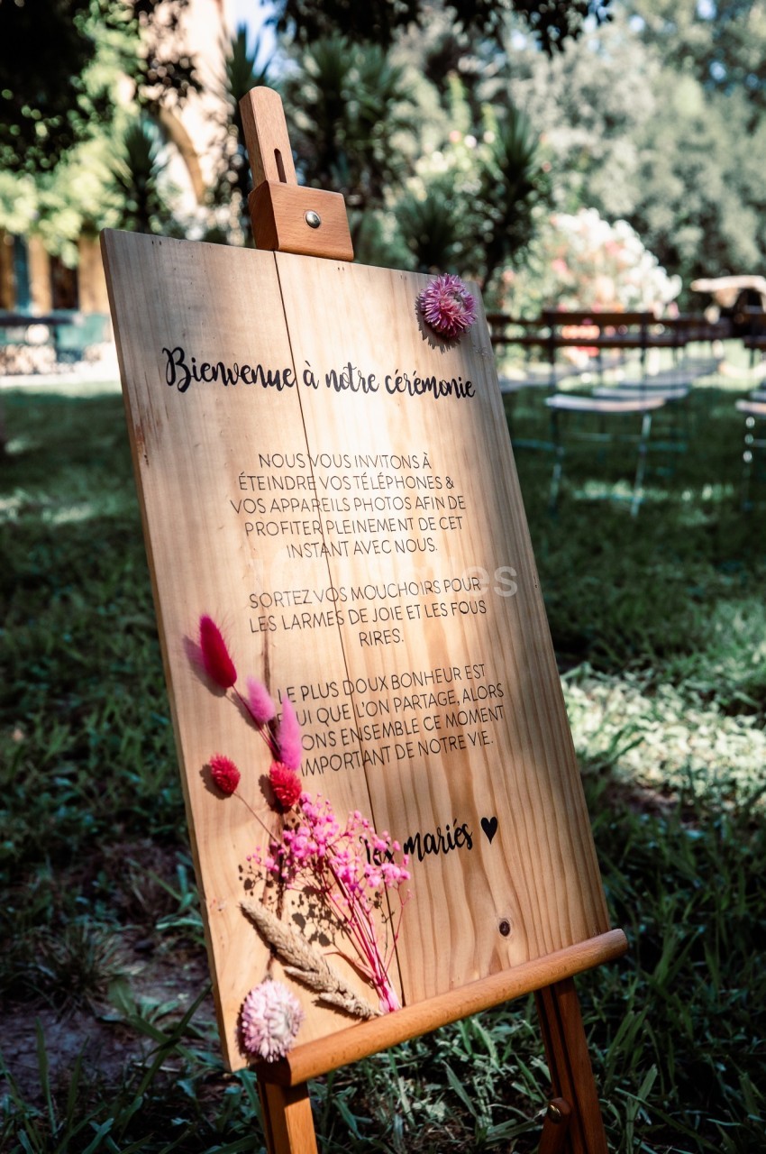 Panneau en bois sur un chevalet, décoré de fleurs séchées, avec un message de bienvenue pour une cérémonie en extérieur.