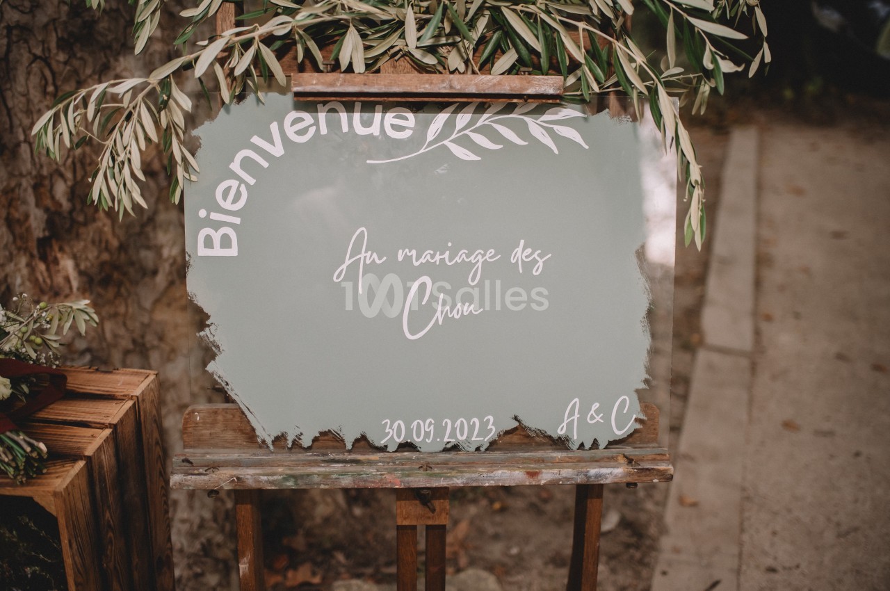 Panneau de bienvenue peint à la main pour un mariage, posé sur un chevalet en bois, entouré de feuillage.