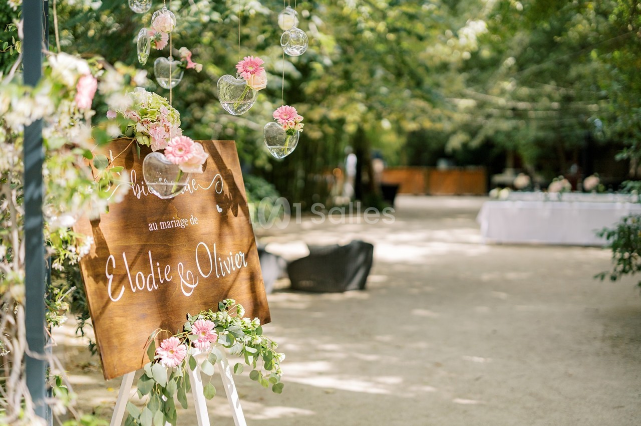 Panneau en bois décoré de fleurs roses annonçant un mariage, installé dans une allée arborée et lumineuse.