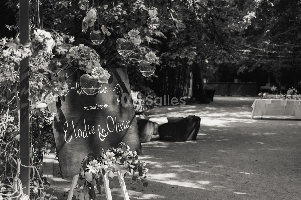 Panneau décoratif annonçant un mariage, entouré de fleurs et suspendu sous des arbres dans un espace extérieur.
