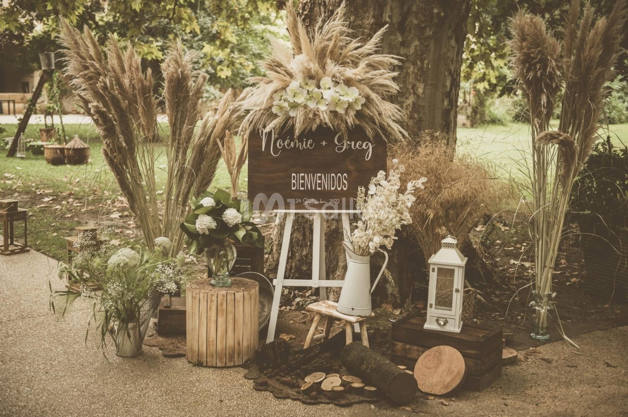 Décoration rustique avec panneau de bienvenue, fleurs séchées, lanternes et éléments en bois sous un arbre.