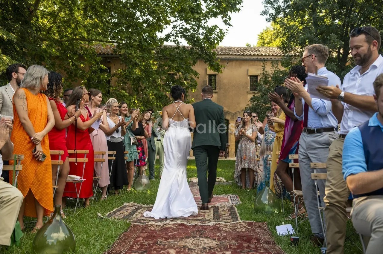 Un couple marche dans une allée extérieure bordée d'invités applaudissant lors d'une cérémonie de mariage.