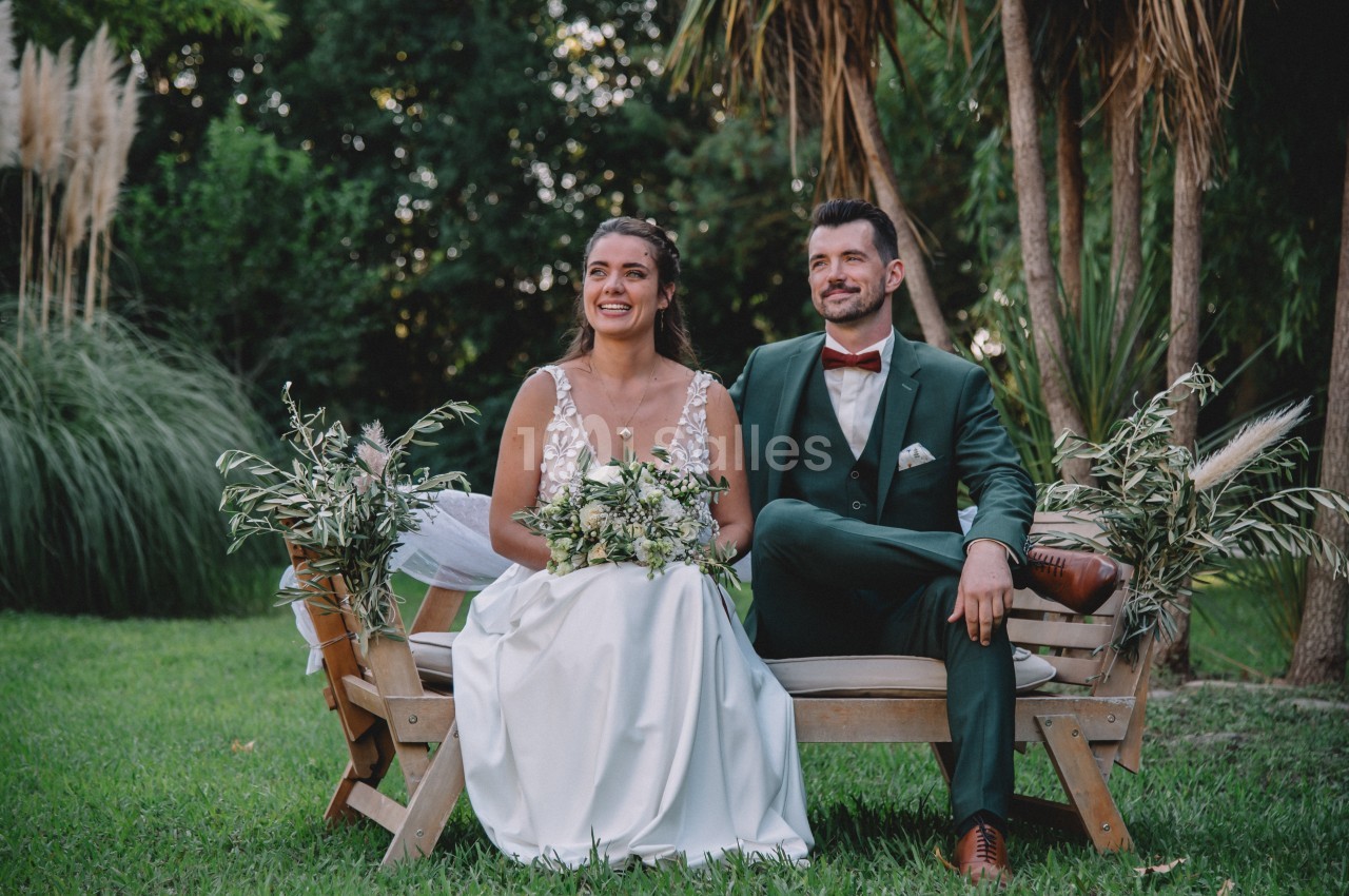 Un couple en tenue de mariage est assis sur un banc en bois dans un jardin verdoyant.