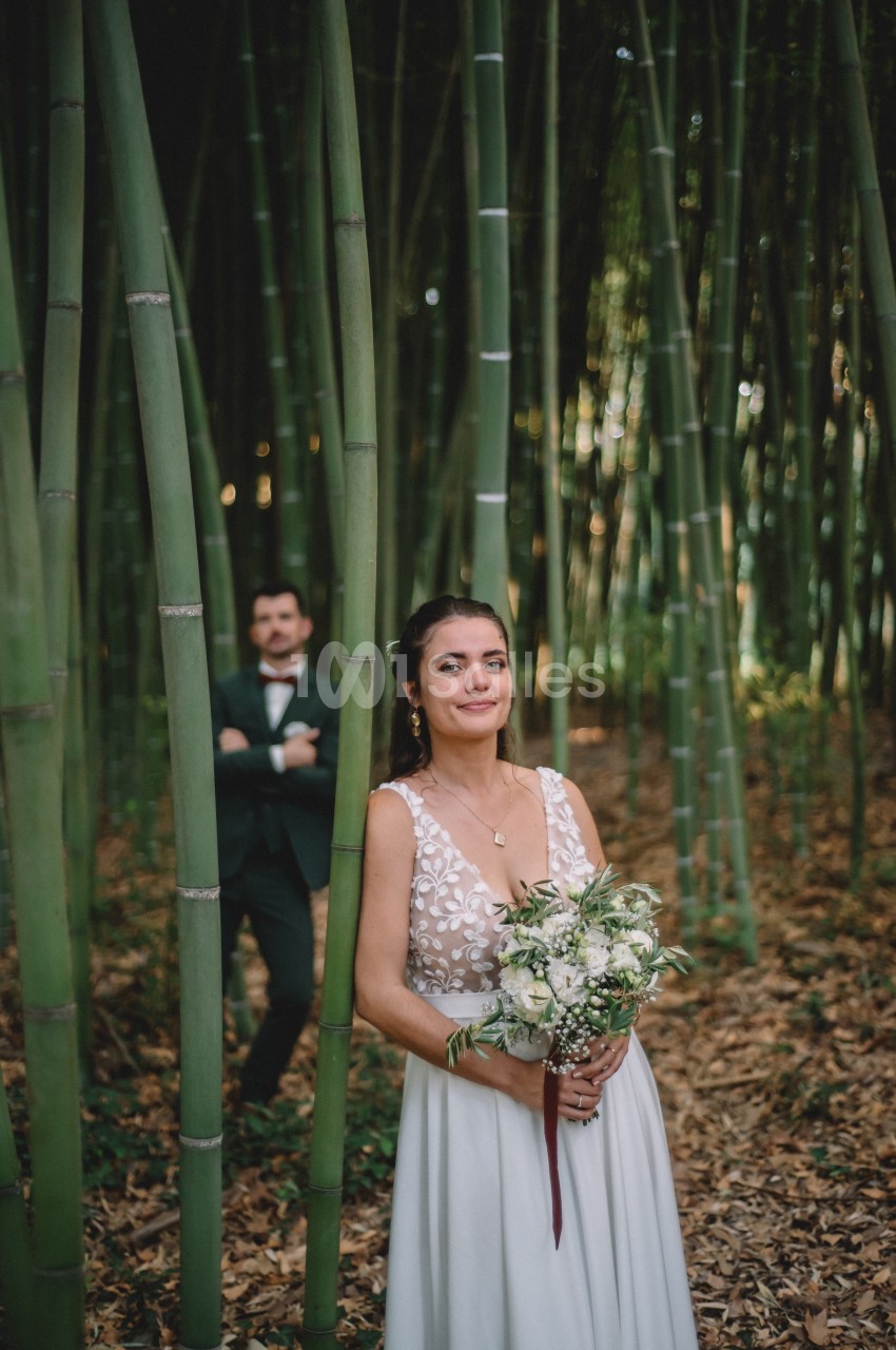 Une femme en robe blanche tient un bouquet dans une forêt de bambous, un homme en costume se tient en arrière-plan.