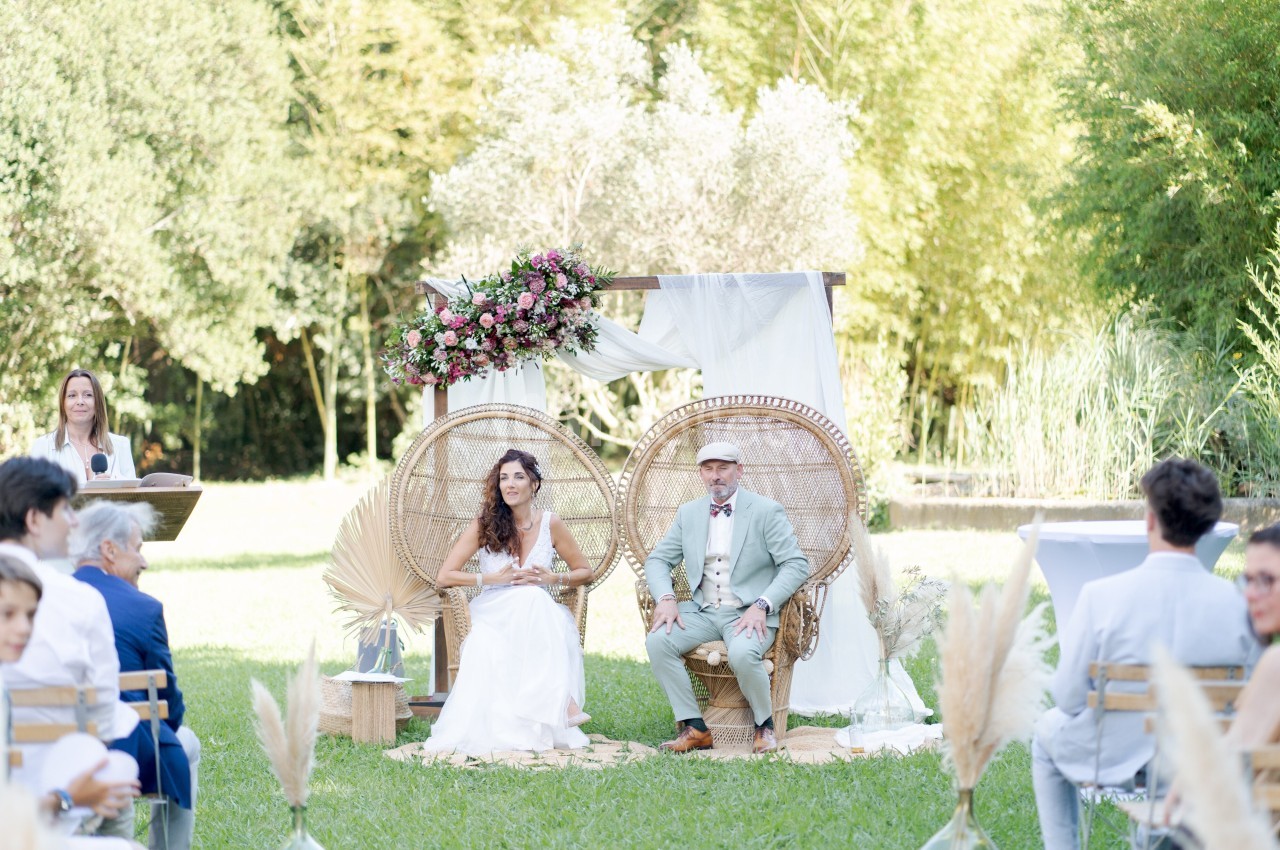 Un couple assis sur des fauteuils en rotin lors d'une cérémonie en plein air, entouré de décorations florales et végétales.