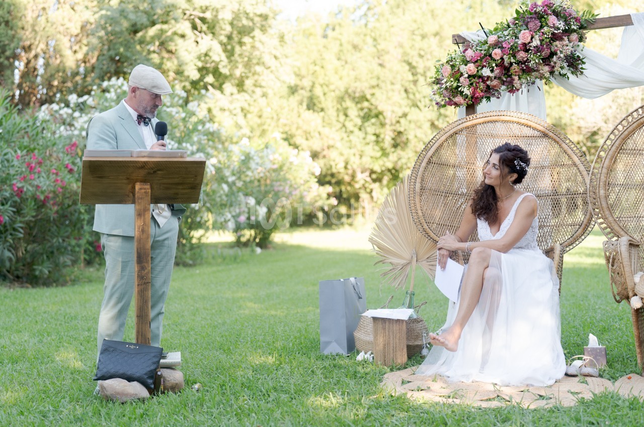 Un homme en costume parle à un micro devant une femme en robe blanche assise sur une chaise en osier dans un jardin fleuri.