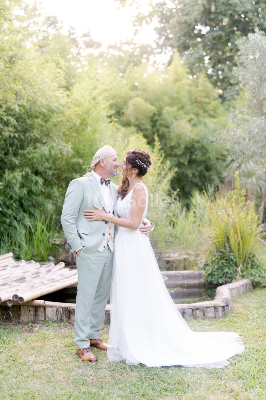 Un couple en tenue de mariage se tient debout dans un jardin verdoyant près d'un petit pont en bois.
